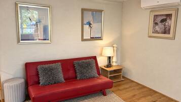 A red sofa with grey cushions, a small side table with a lamp, and framed artwork on the walls.