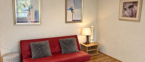 A red sofa with grey cushions, a small side table with a lamp, and framed artwork on the walls.