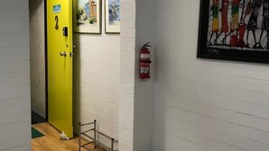 A hallway with a yellow door, a fire extinguisher, framed pictures on the wall, and a green bag on the floor. - Santa Monica apartment (Carnegie)