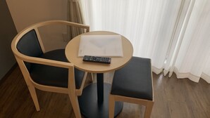Une petite table ronde avec une télécommande et un carnet, deux chaises avec des coussins foncés, et une fenêtre avec des rideaux.