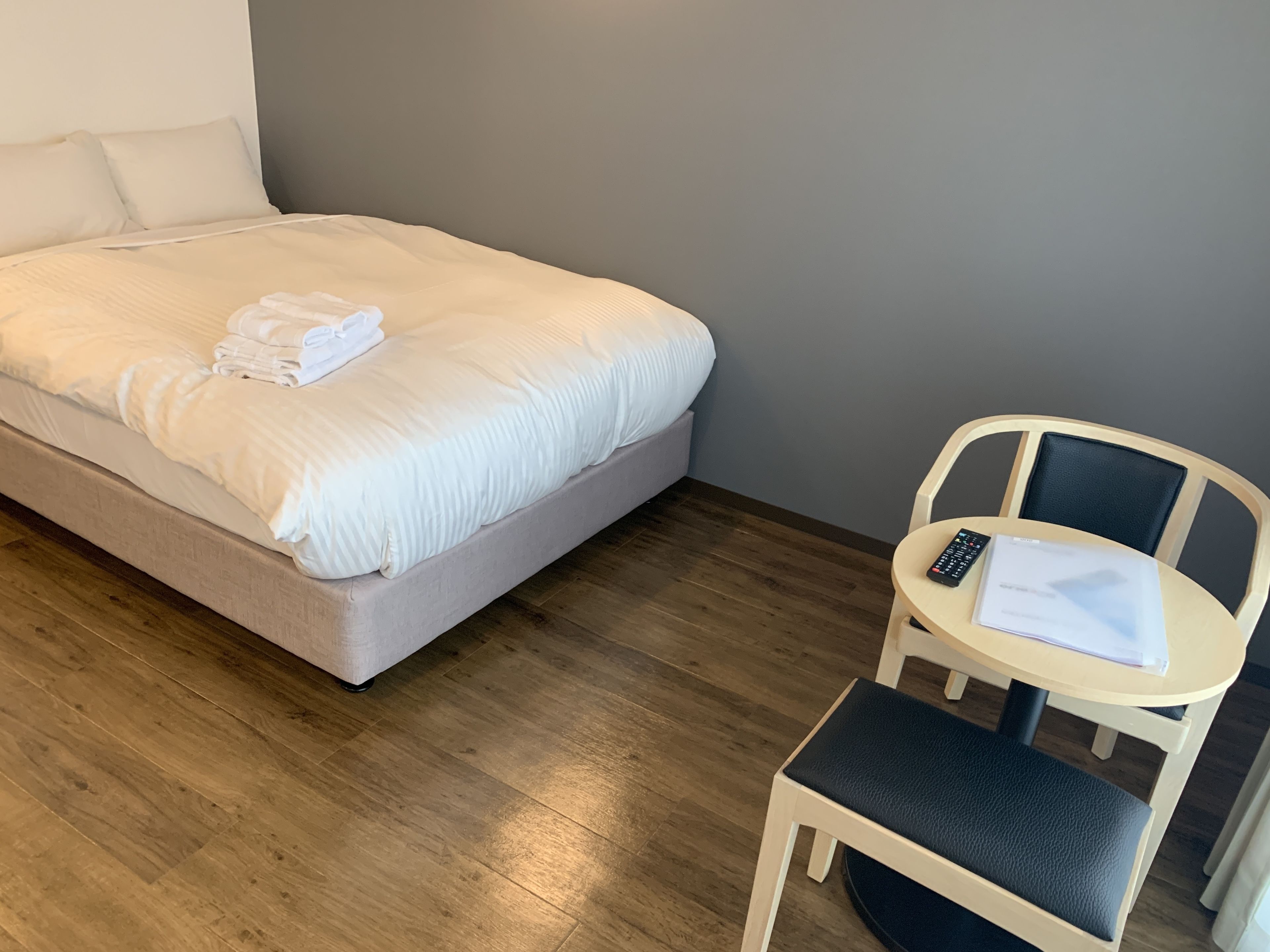 A neatly made bed with white linens and a folded towel, a small round table with a remote control and a booklet, and a chair with a cushion.