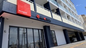 Un bâtiment moderne abritant un magasin M. Kinjo au rez-de-chaussée, de grandes fenêtres en verre et un espace de stationnement délimité par des lignes jaunes.