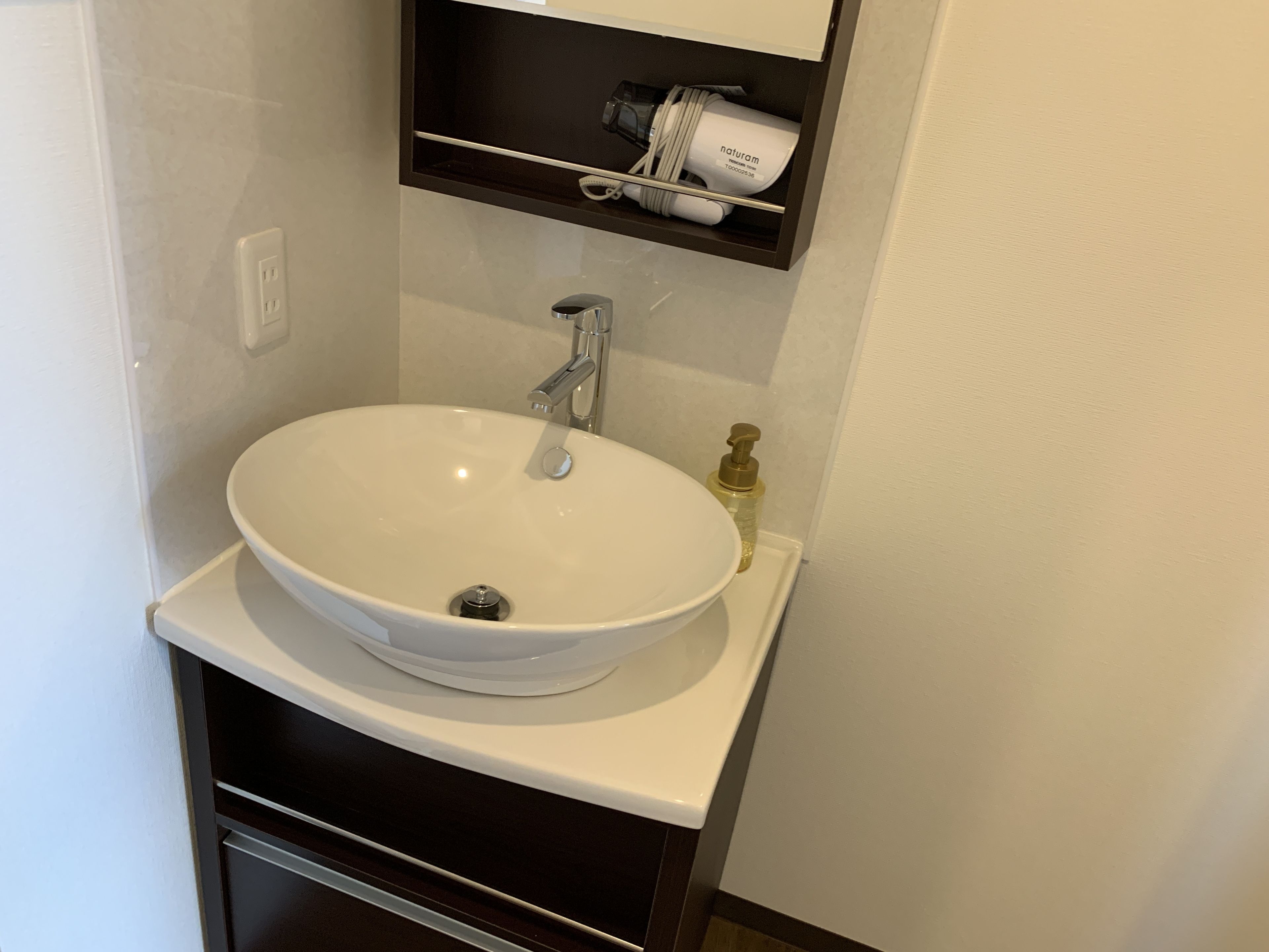 A white bathroom sink with a chrome faucet, a soap dispenser, and a shelf above holding a hairdryer and a bottle.