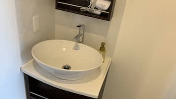 Un lavabo blanc dans la salle de bain, doté d’un robinet chromé, d’un distributeur de savon et d’une étagère au-dessus, sur laquelle sont posés un sèche-cheveux et une bouteille.
