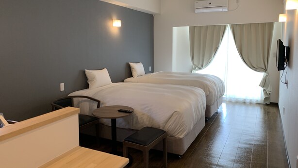 Une chambre d’hôtel avec deux lits, une petite table, un banc et un téléviseur fixé au mur.