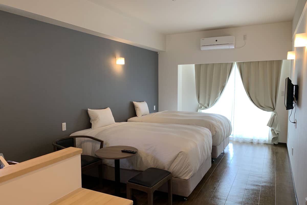 A hotel room with two beds, a small table, a bench, and a TV mounted on the wall.