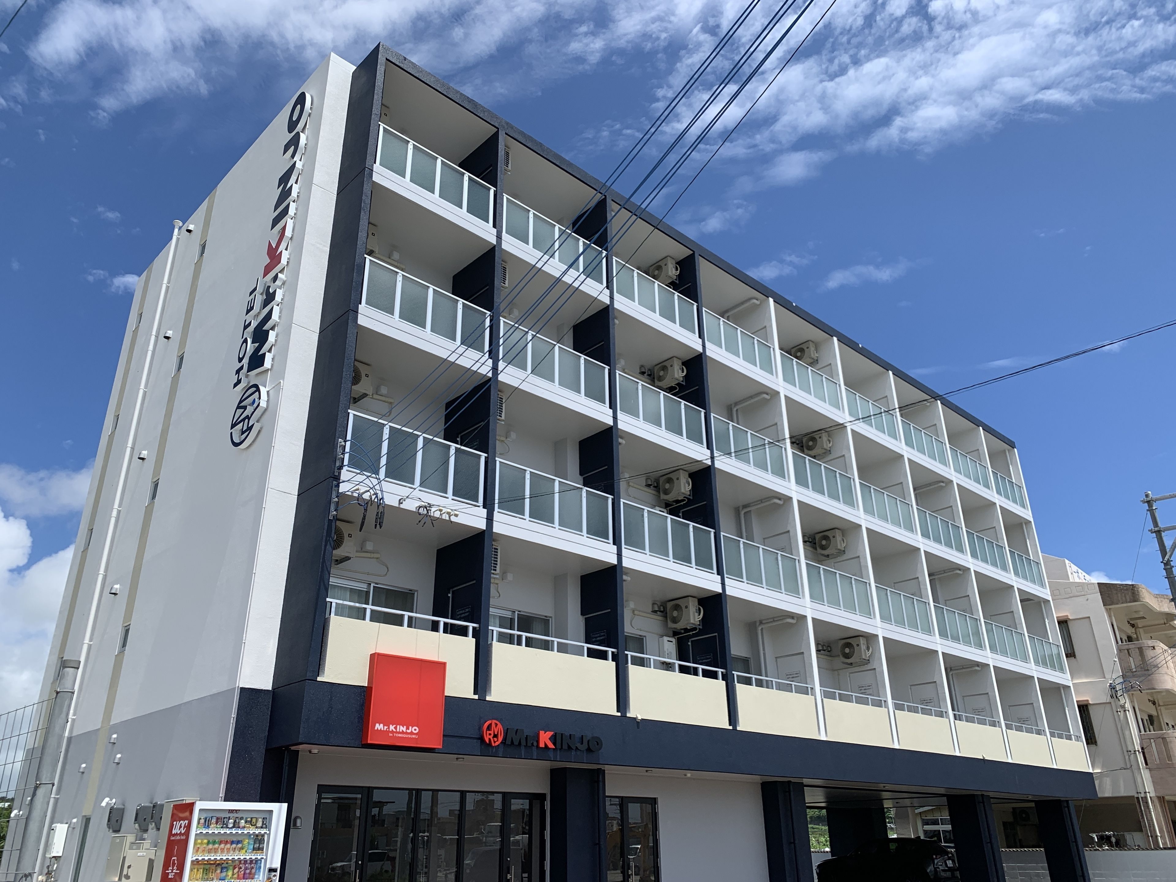 A multi-story hotel with a modern design, featuring balconies on each floor and a prominent Mr. Kinjo sign at the ground level.