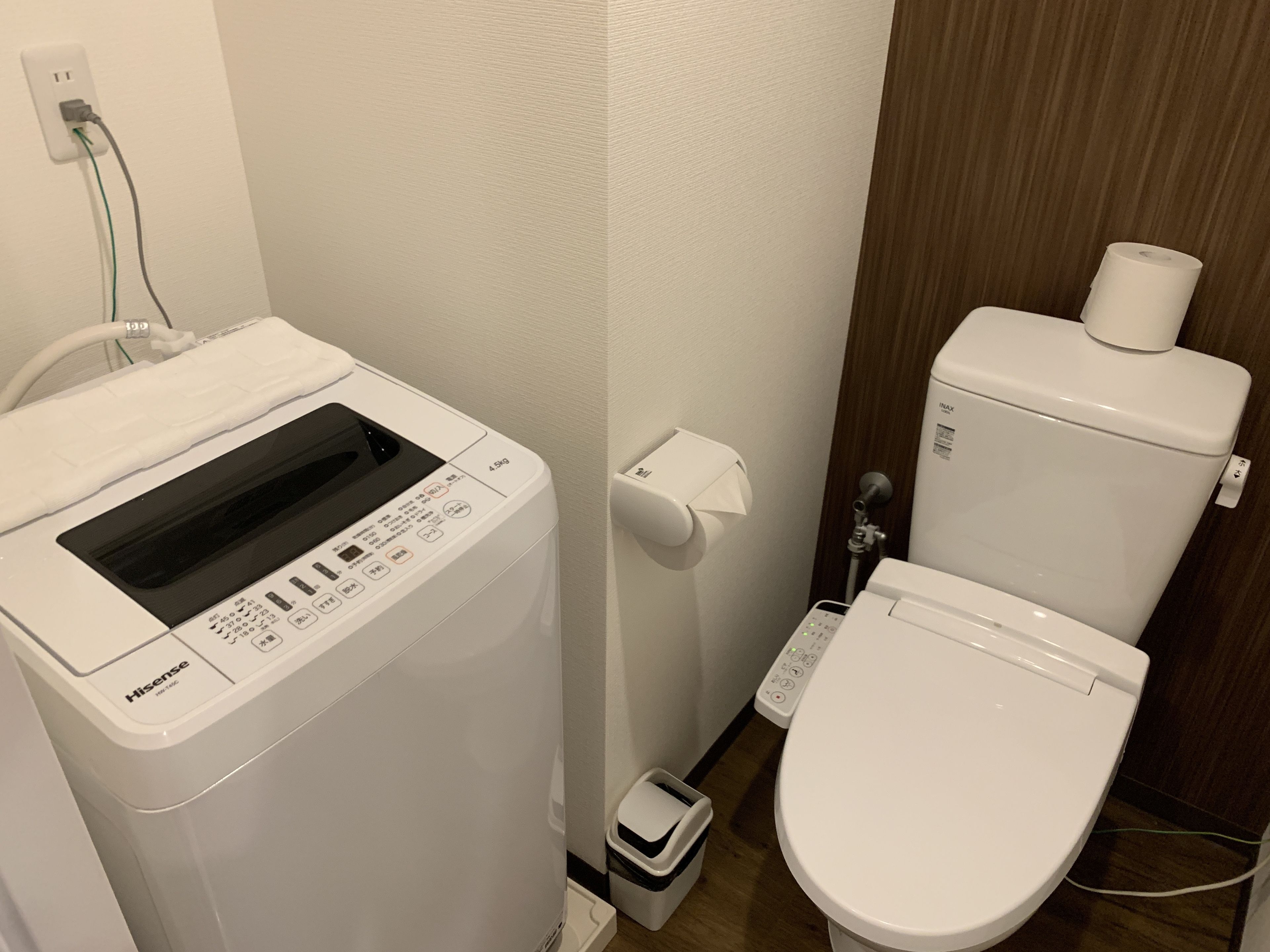 A compact bathroom with a white Hisense washing machine, a toilet, and a paper towel dispenser.