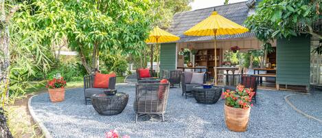 An outdoor seating area with wicker furniture and yellow umbrellas, surrounded by greenery and potted plants.