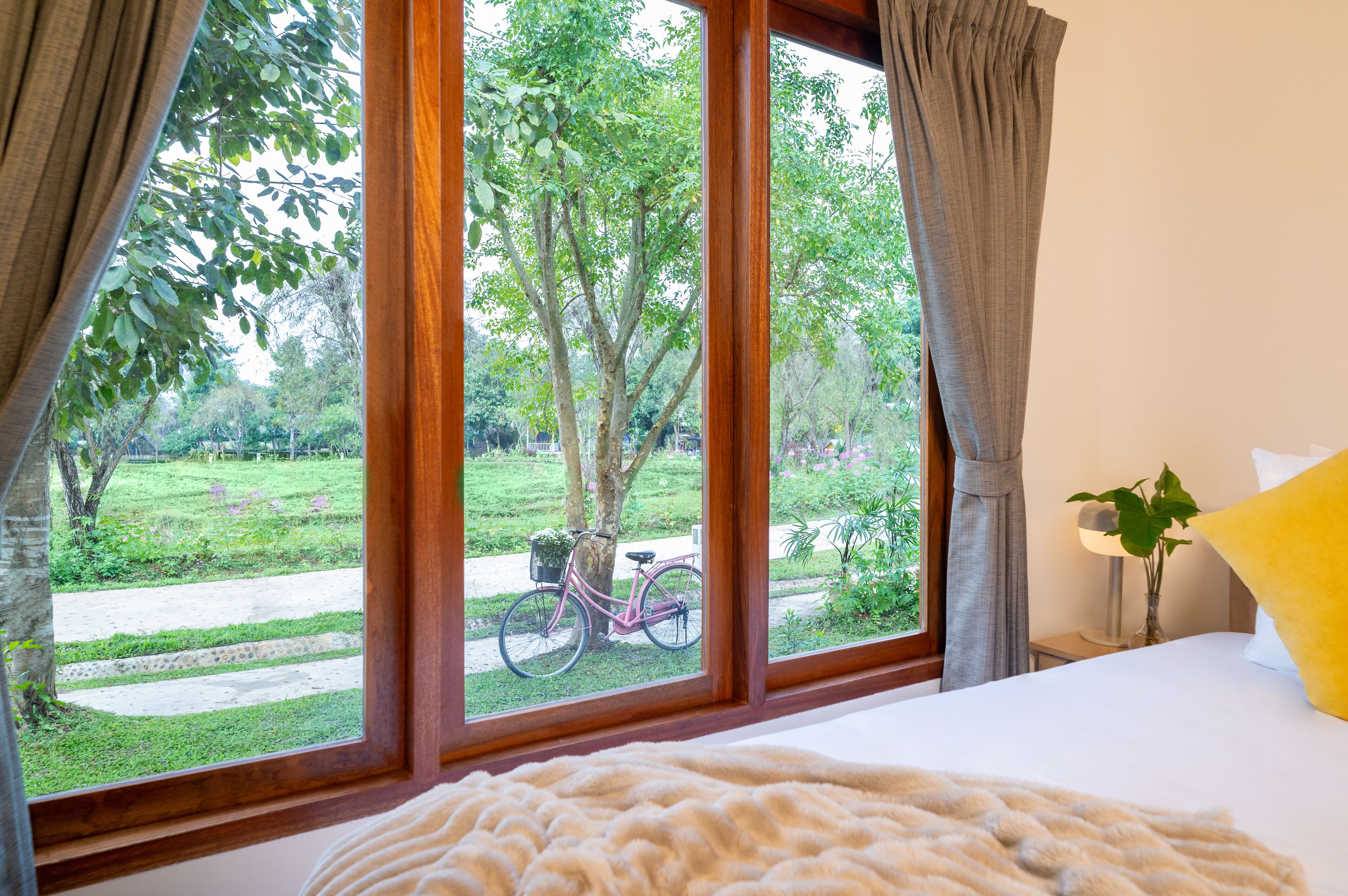 Une chambre avec une grande fenêtre, un lit recouvert d’une couverture blanche, un oreiller jaune et un vélo à l’extérieur.
