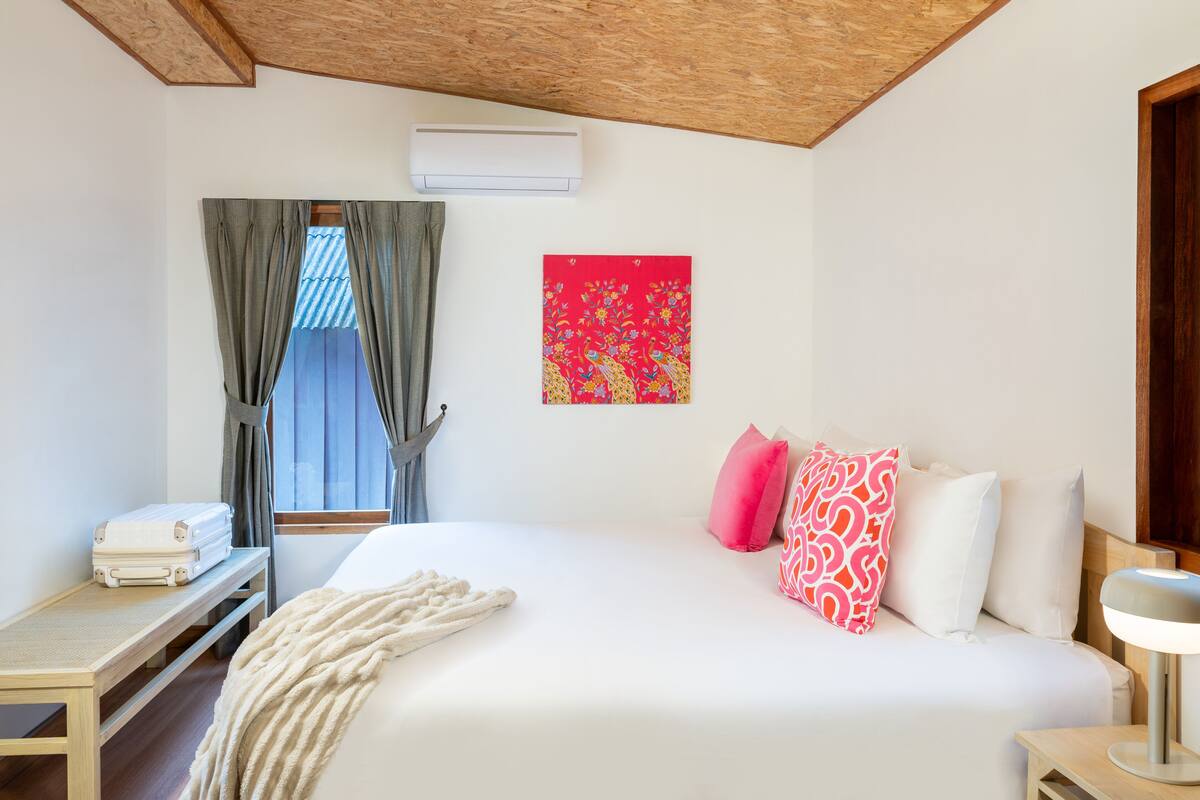 A hotel room with a bed, a bedside table with a lamp, a window with curtains, an air conditioning unit, and a wall-mounted artwork.
