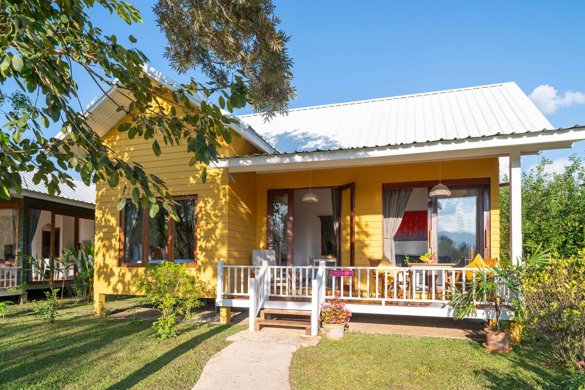 A yellow house with a white porch, a small garden, and a pathway leading to the entrance.