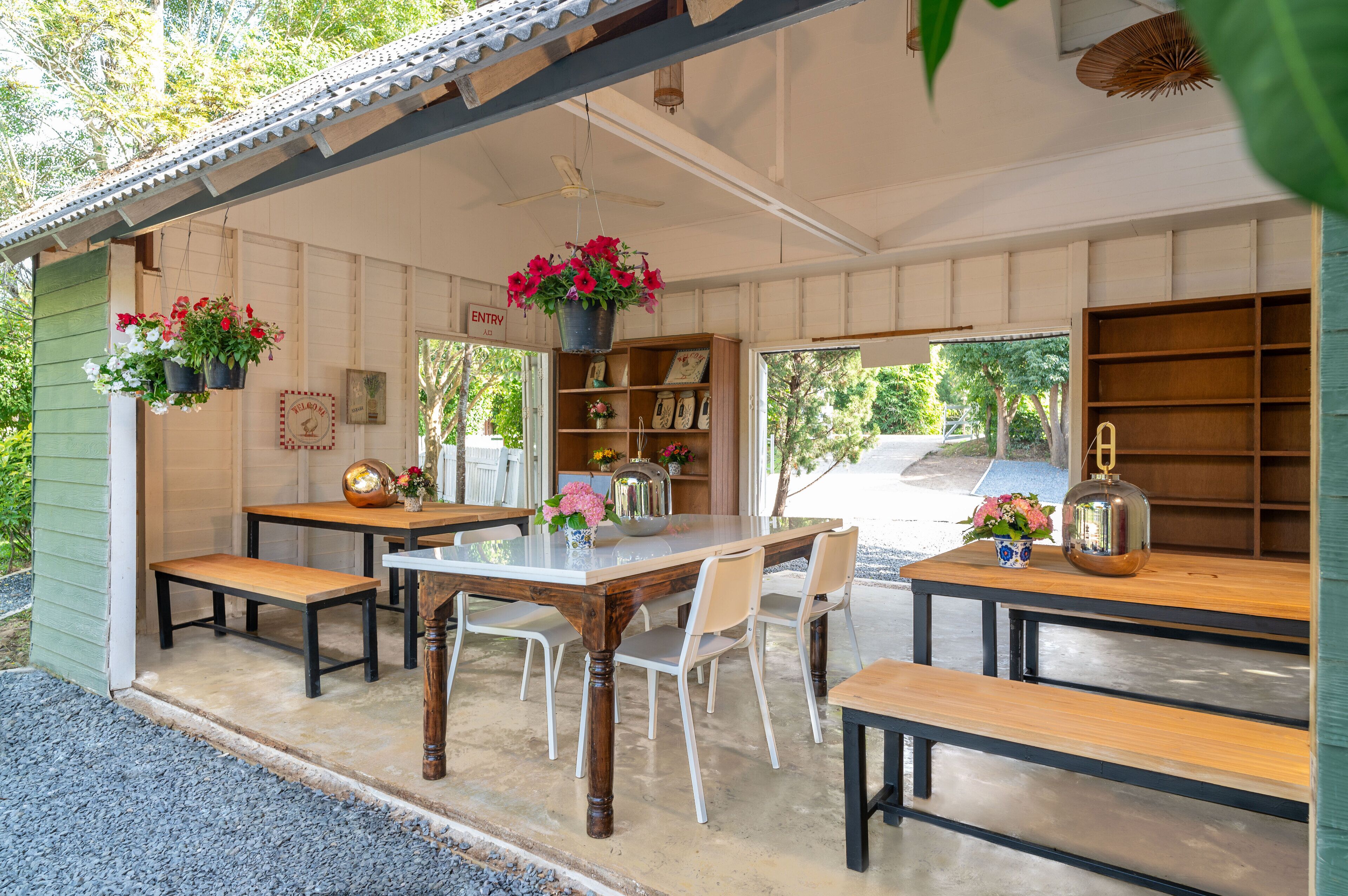A covered outdoor dining area with tables and benches, surrounded by a wooden structure with hanging flower baskets and a bookshelf.