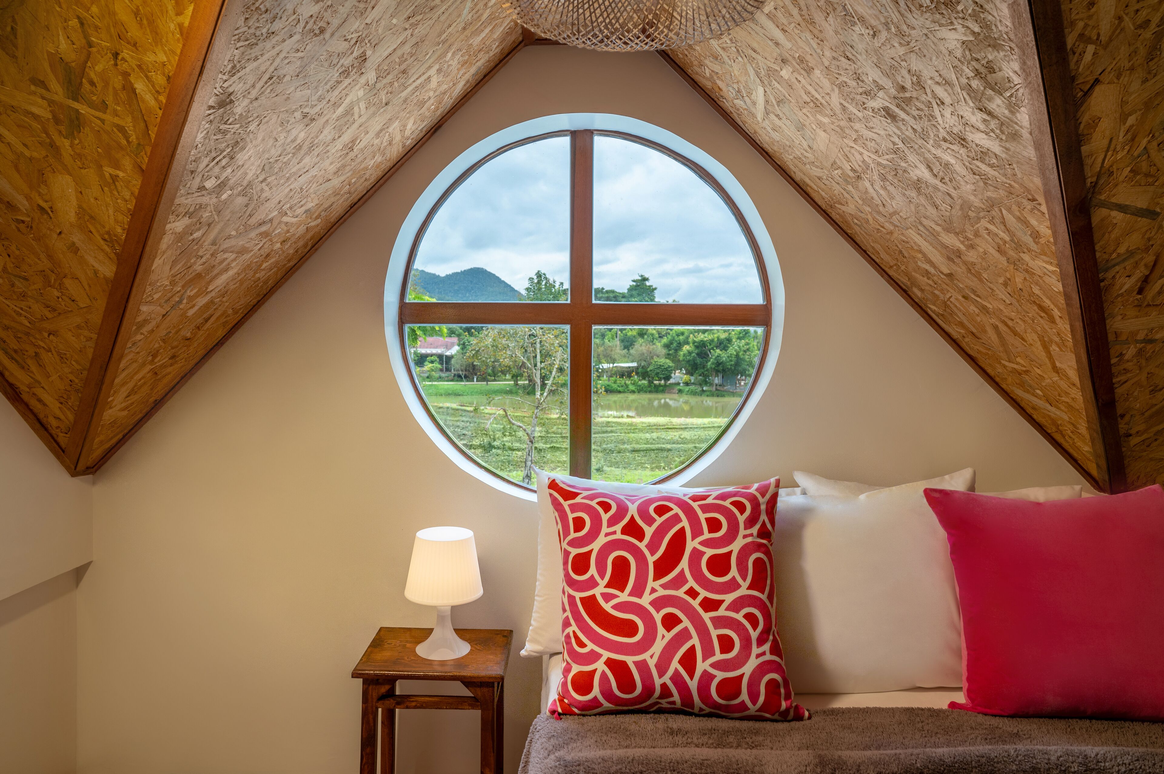 A bedroom with a large circular window, a bed with red and white pillows, and a small bedside table with a lamp.