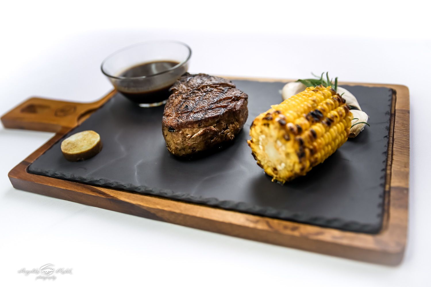 Een steak en gegrilde maiskolf op een zwarte leisteenplank met een klein schaaltje saus.