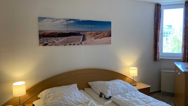 A hotel room with a single bed, two bedside lamps, a nightstand with a phone, and a wall-mounted picture of a beach scene. - House Hayer - House Hayer Whg 10 (Sankt Peter-Ording)