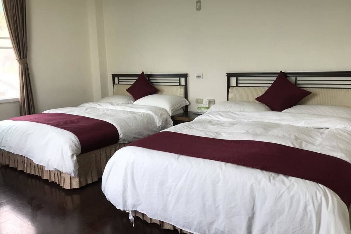 Two beds with white linens and maroon throw pillows, a wooden floor, and a window with curtains.