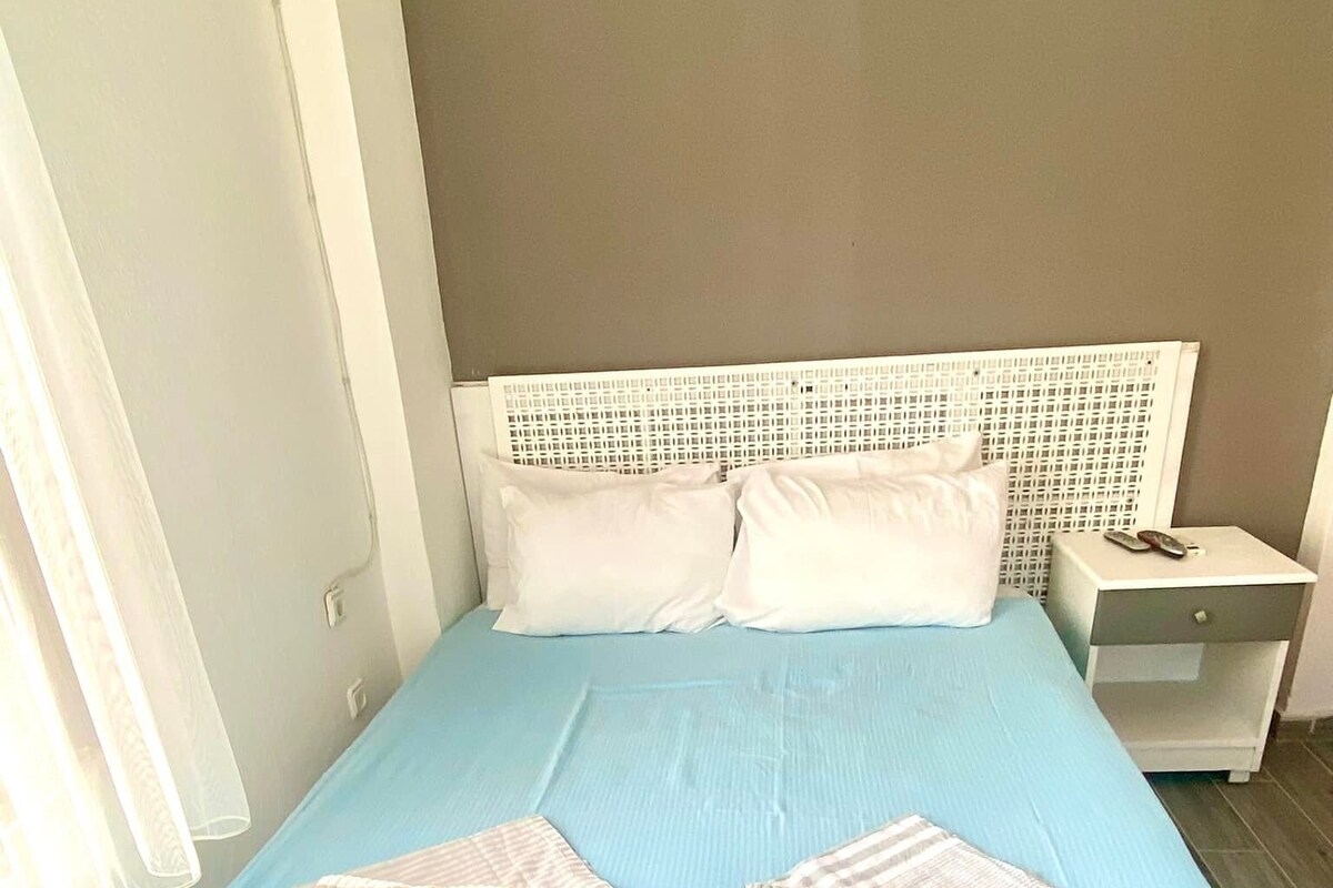 A bed with a blue bedspread and two white pillows, a white headboard, and a bedside table with a remote control.