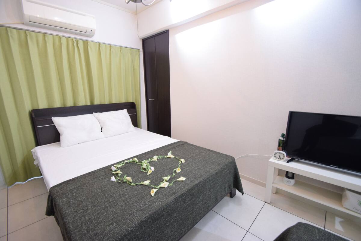 A neatly made bed with a heart-shaped decoration, a television on a stand, and an air conditioning unit above the bed.