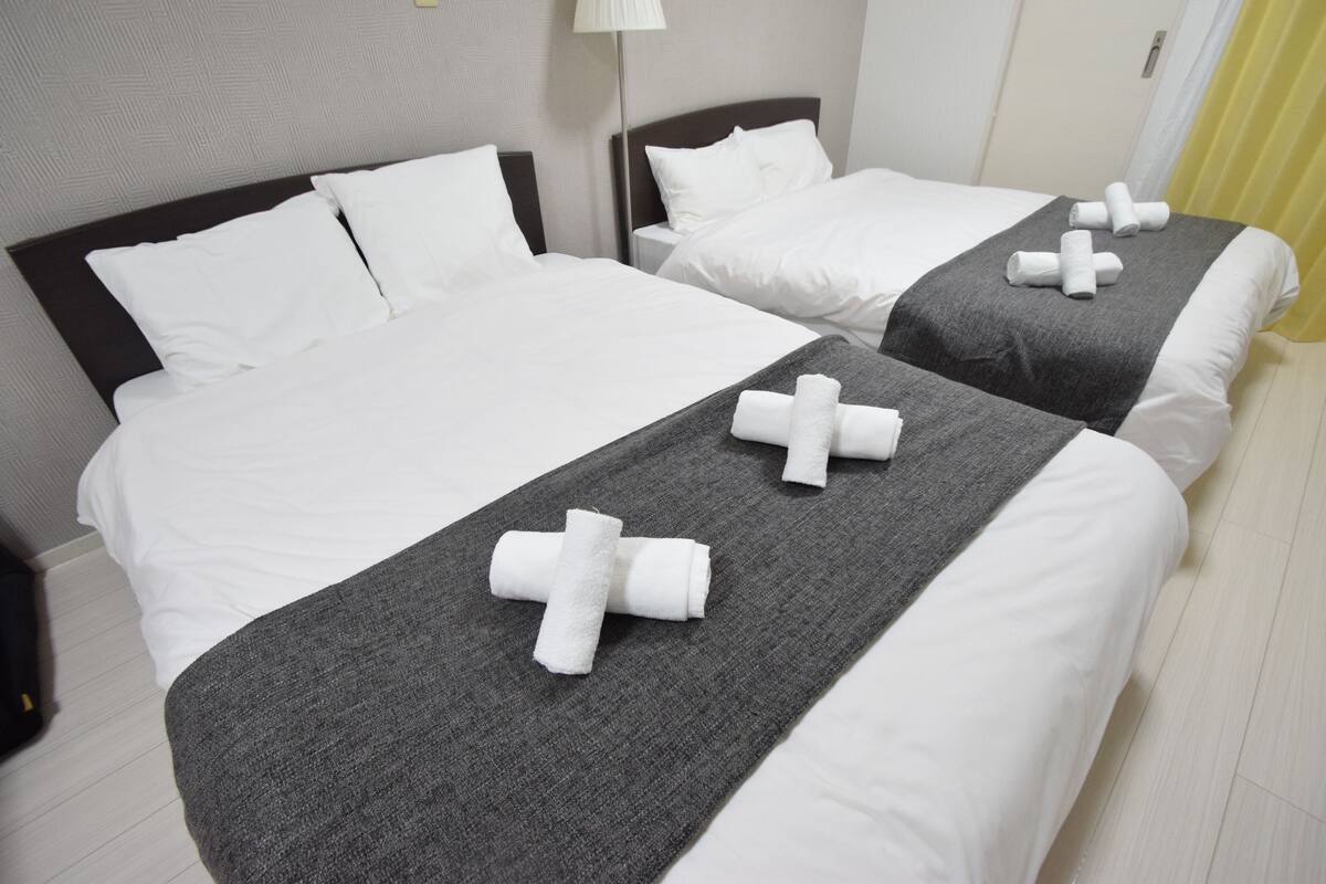 A hotel room with two beds, each with white linens and a grey blanket. There are towels arranged on the beds.
