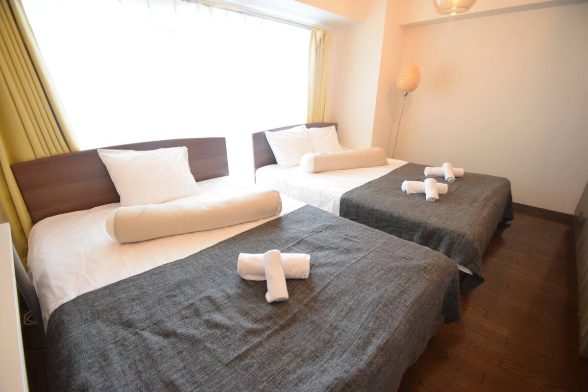 A hotel room with two beds, each with a grey bedspread and white pillows. There is a window with curtains, a bedside lamp, and a wooden floor.