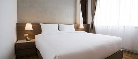 A hotel room with a large bed, wooden headboard, two bedside tables with lamps, and a view of the outdoors through sheer curtains.