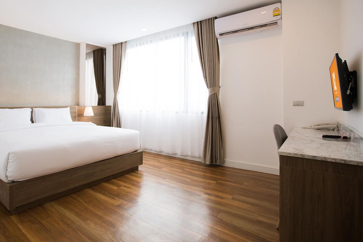 A hotel room with a large bed, wooden flooring, a desk with a chair, a TV mounted on the wall, and a window with curtains.