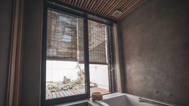A bathroom with a bathtub, a window with blinds, and a view of a garden. - Modern Machiya MIBU (Kyoto)