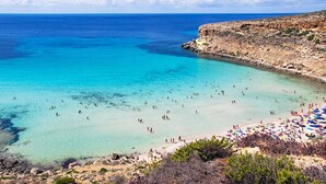 Una playa con aguas turquesas cristalinas, acantilados rocosos y una orilla arenosa con sombrillas.