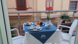 Un balcón con una mesa lista para dos personas, con mantel azul que lleva un logotipo, sillas blancas y una planta en maceta con flores rojas.