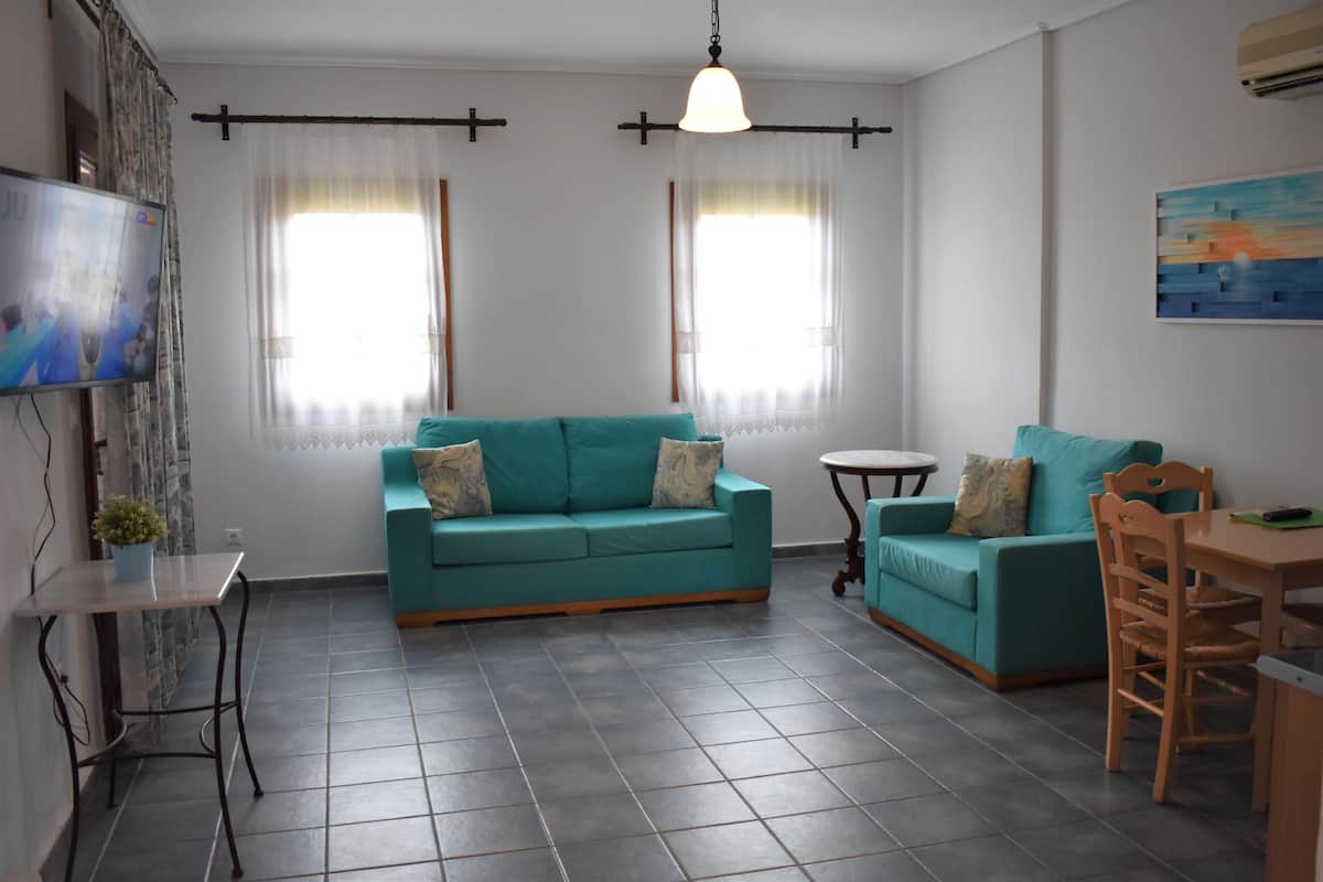 A living room with two teal sofas, a small round table, a wooden chair, and a TV mounted on the wall.