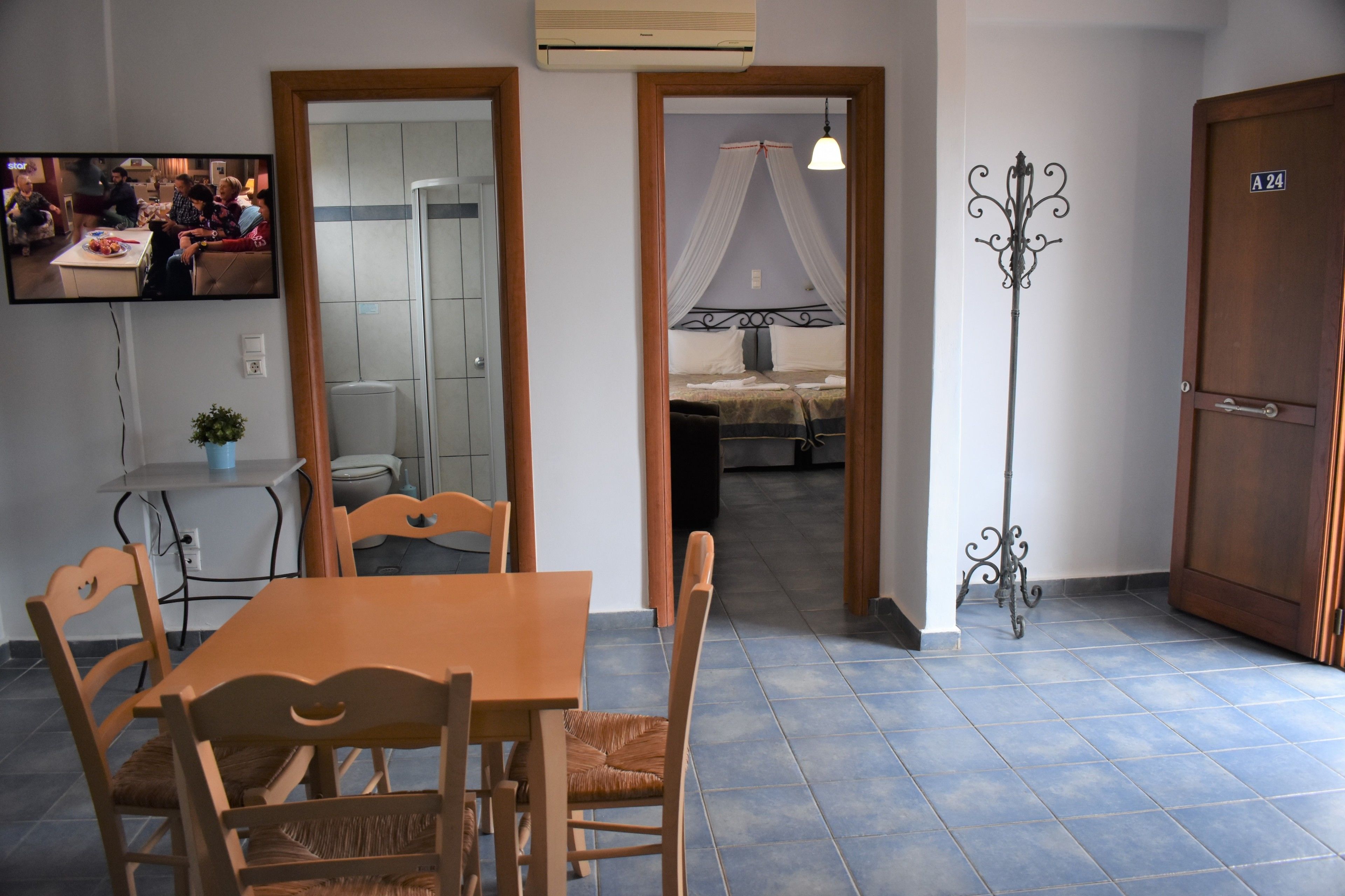 A dining area with a wooden table and chairs, a TV mounted on the wall, and a bed with a canopy in the background.