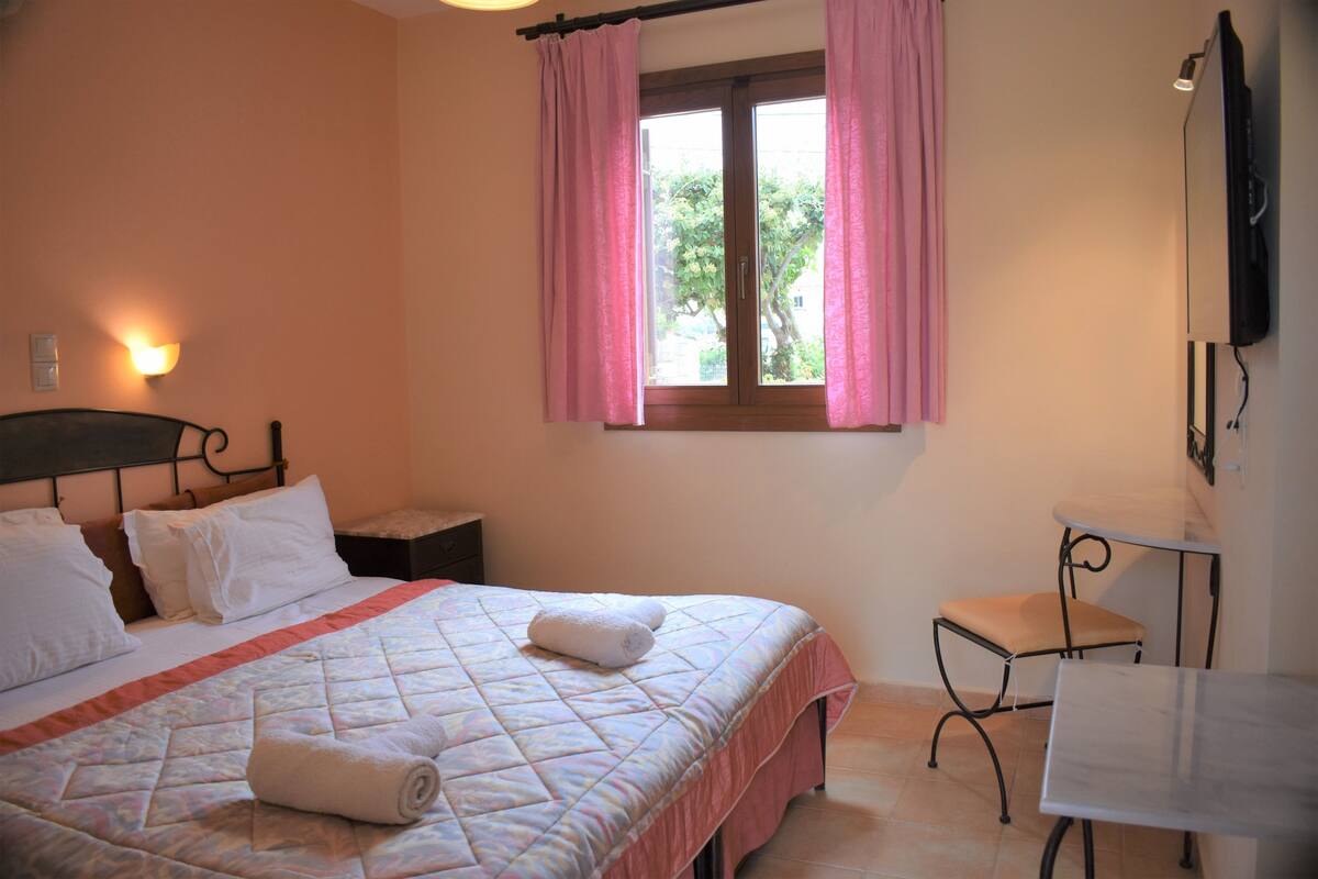A neatly made bed with a quilt, pillows, and rolled towels. There is a small bedside table and a chair next to the bed. A window with pink curtains is visible, and a flat-screen TV is mounted on the wall.