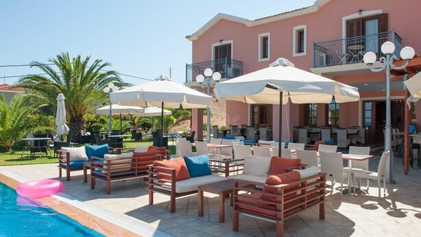 A poolside area with lounge chairs and umbrellas, a pink building with balconies, and a clear sky. - Melidron Hotel (Kefalonia)