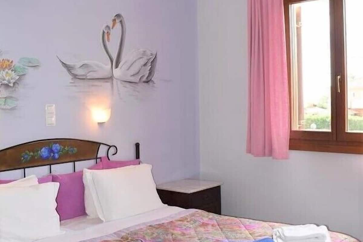 A bedroom with a bed, pillows, a nightstand, a wall-mounted light, and a window with curtains.