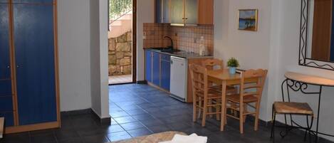 A compact room with a bed, a small dining table with chairs, a kitchenette with blue cabinets, and a stone wall visible through an open door.