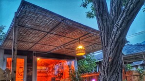 Una casa de madera rústica con techo de paja, un gran árbol en primer plano y un interior acogedor con sofá y mesa.