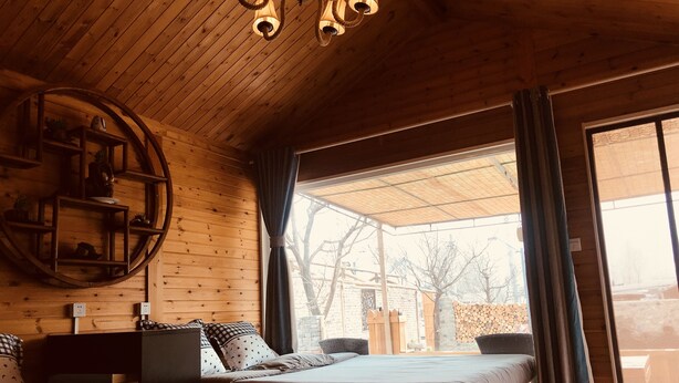 Una habitación acogedora con paredes de madera y techo inclinado, que incluye dos camas, un estante circular y un amplio ventanal con cortinas.