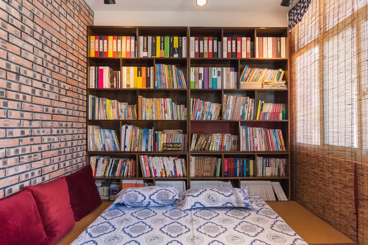 A room with a brick wall, a large bookshelf filled with books, a bed with patterned bedding, and red cushions.