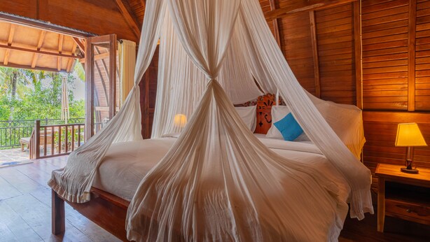 A wooden four-poster bed with a canopy, a nightstand with a lamp, and a view of the outdoors. - Sukawati Inn (Penida Island)