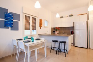 A modern kitchen with white cabinets, a central island with stools, a refrigerator, and a window with blinds. - Trendy with Balcony on Sheinkin by FeelHome (Tel Aviv)