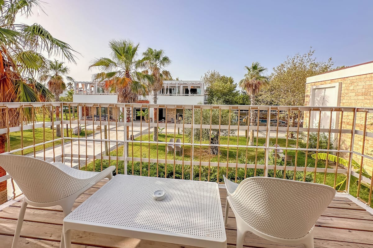 Eine Terrasse mit weißem Metallmöbel, Blick auf einen Innenhof mit Palmen und ein Gebäude im Hintergrund.