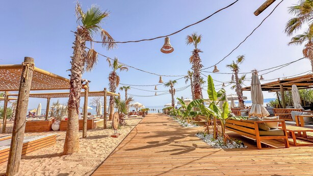 Una terraza de madera que da a una zona de playa con palmeras, bancos de madera y sombrillas.