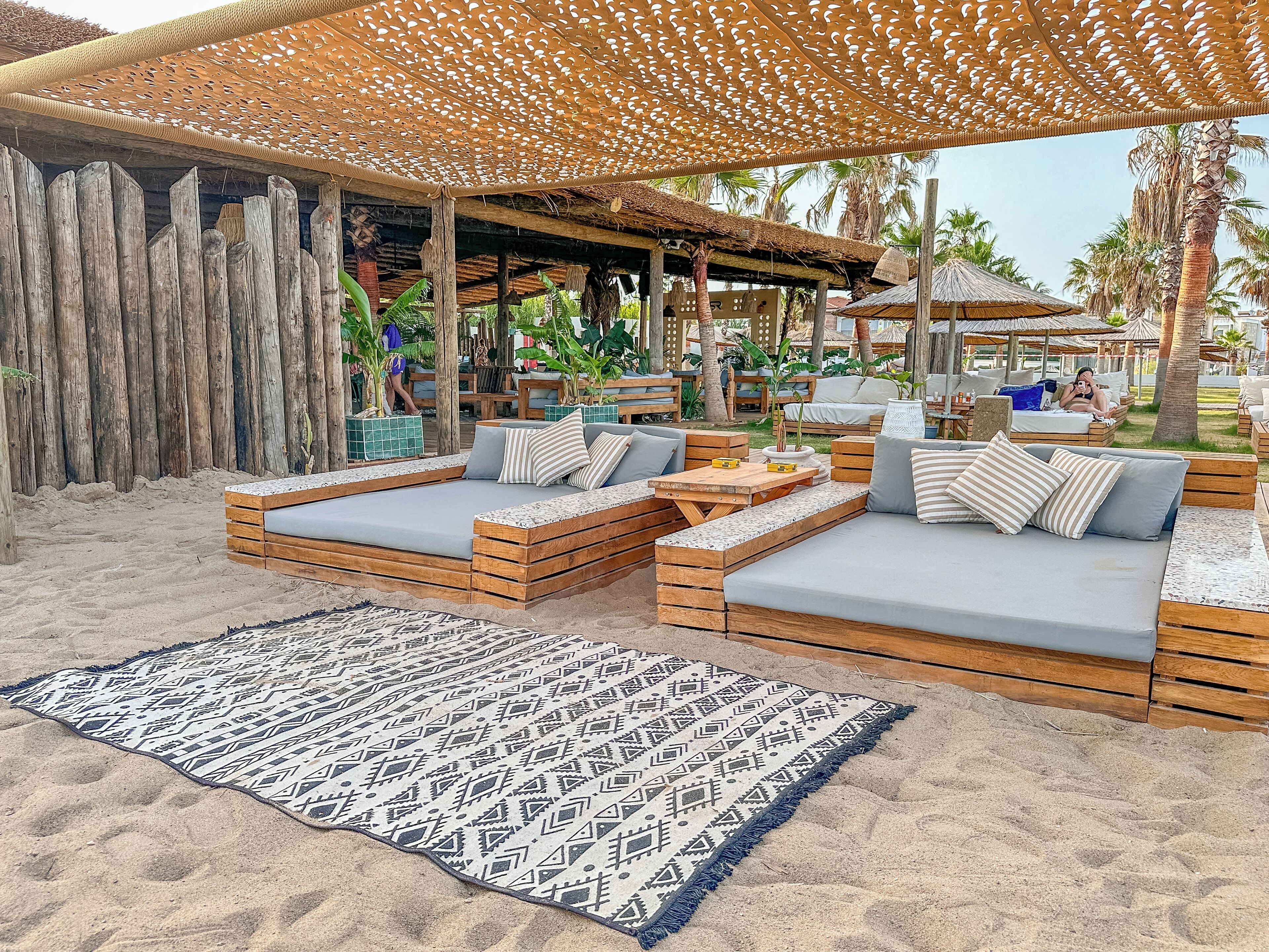 Ein Strandloungebereich mit Holzliegen, einem gemusterten Teppich und einer Struktur mit Reetdach.