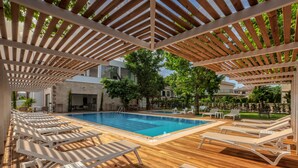 Zona de piscina con terraza de madera, sillas reclinables blancas y una pérgola.