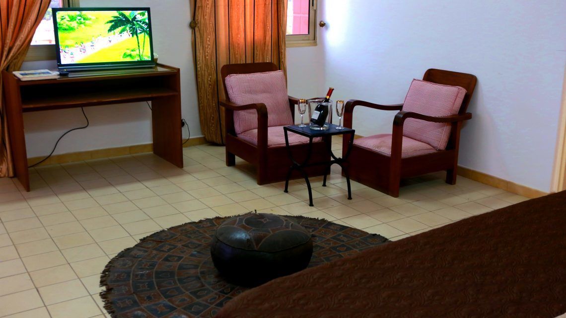 Un salon avec un ordinateur portable posé sur un bureau, deux fauteuils, une petite table avec une bouteille et des verres, et un tapis décoratif.