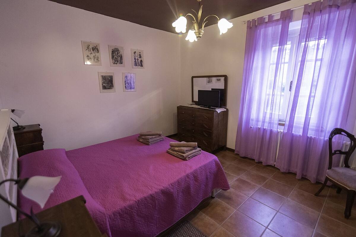Un dormitorio con una cama, un aparador, una silla, un televisor sobre el aparador y una ventana con cortinas moradas.