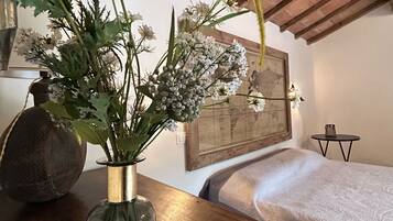 Ein Zimmer mit Holzbalkendecke, einem Bett mit weißer Bettdecke, einem kleinen Tisch mit Blumenvase und einer gerahmten Karte an der Wand.