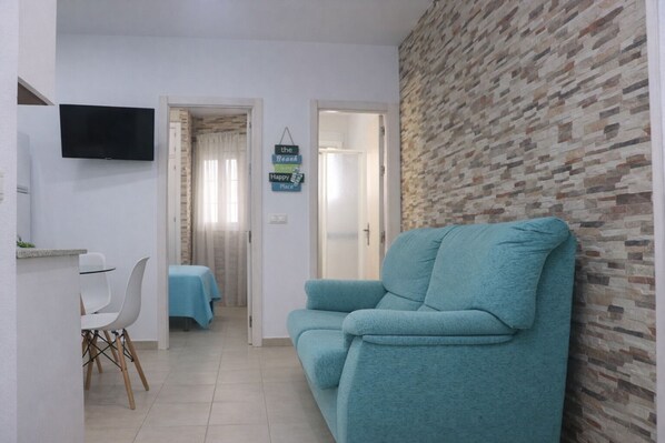 A living room with a teal sofa, a TV mounted on the wall, a dining area with a table and chairs, and a hallway leading to a bedroom with a bed.
