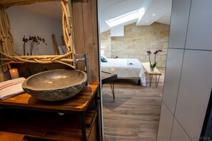 Une salle de bain moderne avec un grand lavabo en pierre, des meubles en bois et, au fond, un lit recouvert d’une courtepointe blanche.